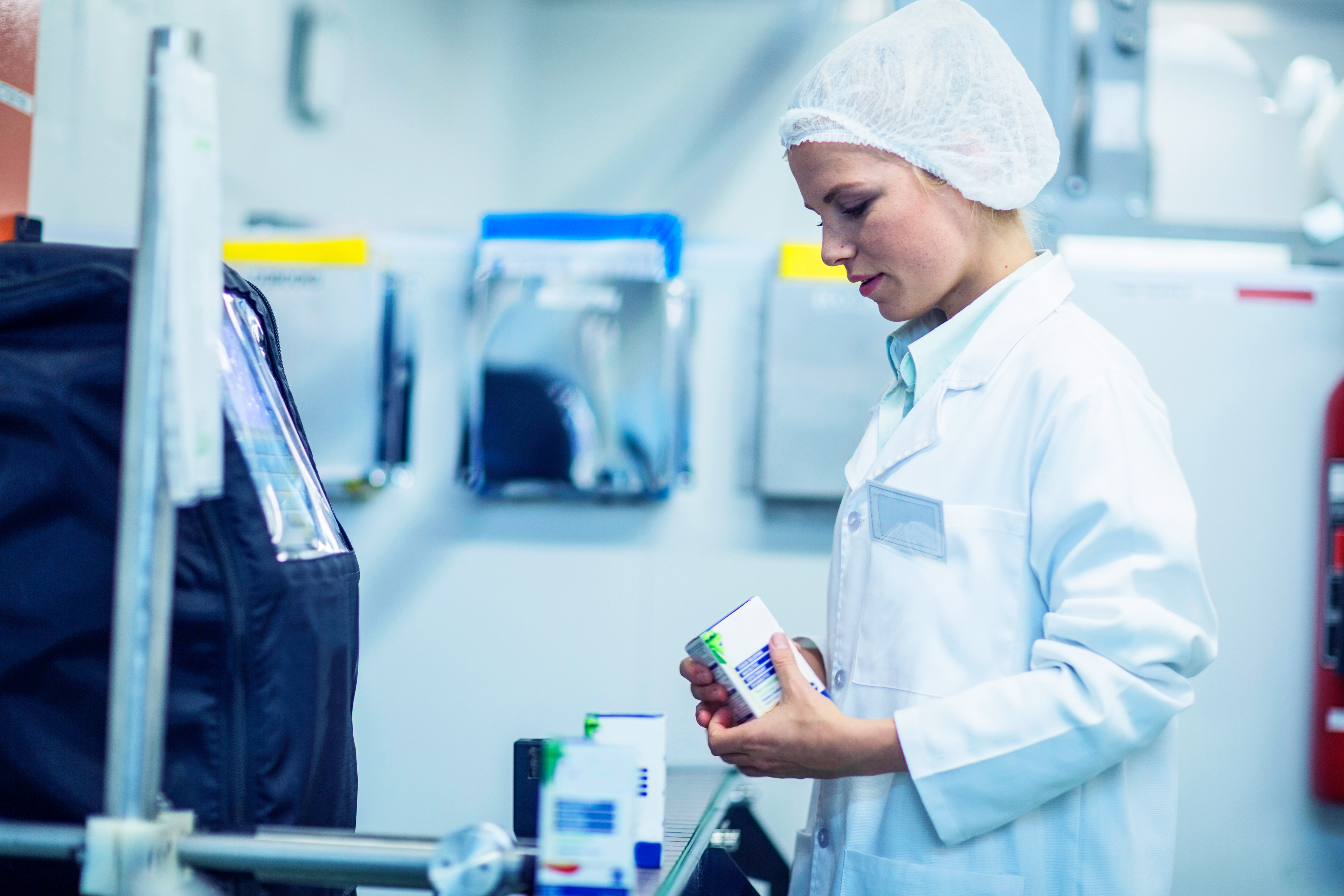 A focused female technician in a lab coat inspects a pharmaceutical product for secondary recovery packaging, ensuring quality control before the packaging launch in a clinical manufacturing setting.