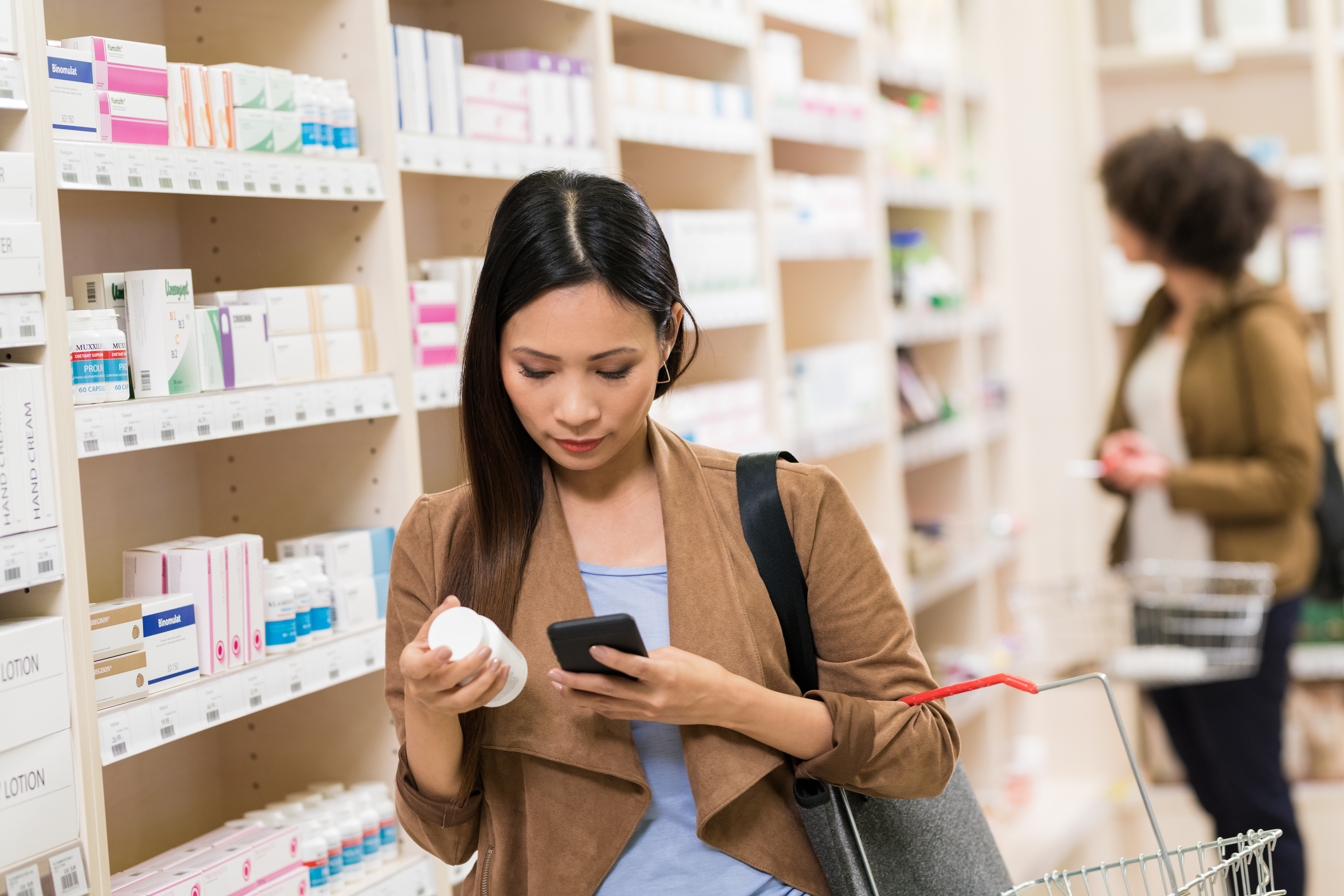 A woman stands in a pharmacy aisle, holding a medication bottle with smart packaging and using her smartphone to scan or check information about the product.