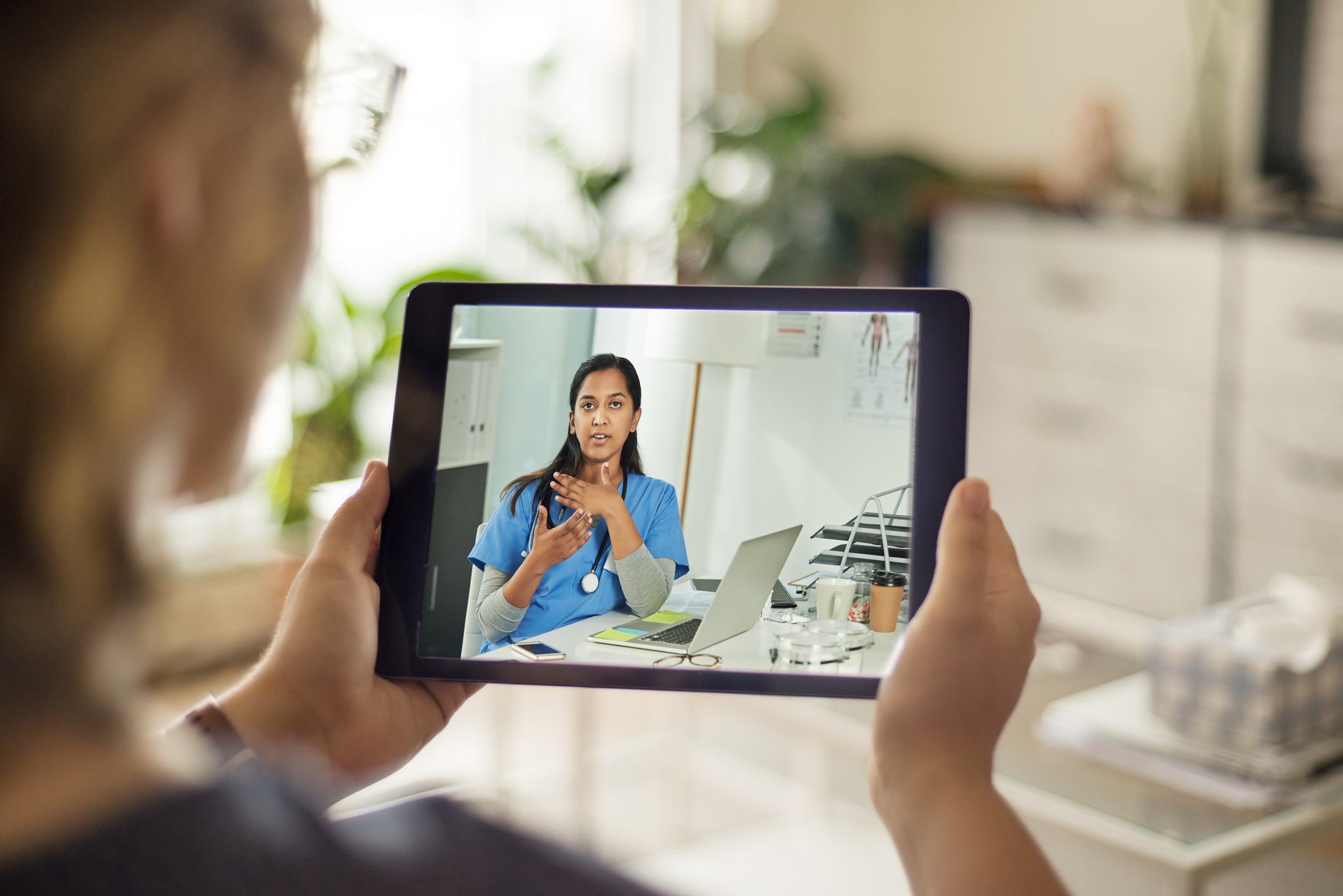 A person is holding a tablet showing a video call with a healthcare professional, who appears to be giving an explanation or consultation. The setting suggests a discussion about pharmaceutical packaging or medication, indicated by the professional demeanor and the office backdrop.