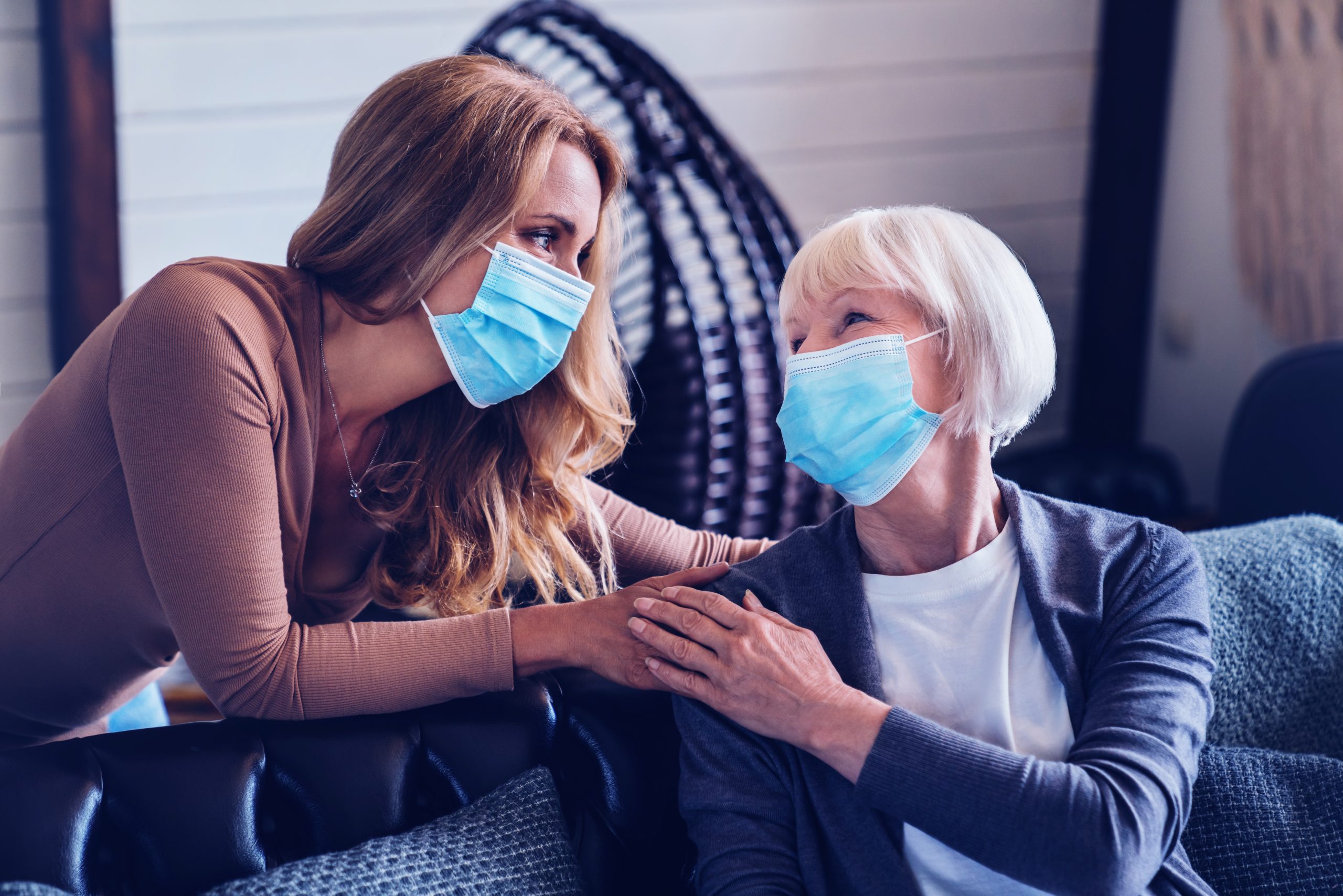 Two women wearing medical masks are engaged in a comforting conversation, with the younger one gently touching the elder's shoulder. The elder looks reassured, possibly discussing something sensitive like pharmaceutical packaging for her medication during a challenging time.