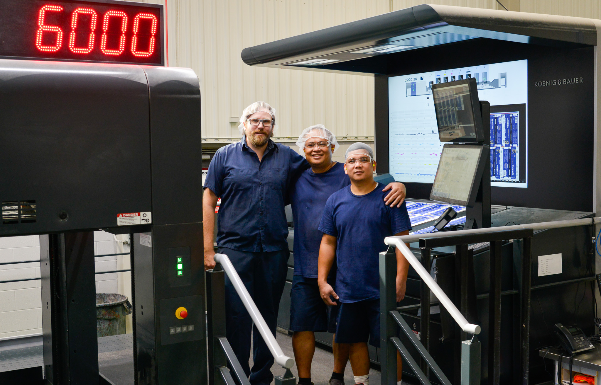 Three workers wearing blue shirts, safety glasses, and hairnets stand together, smiling beside the advanced Koenig & Bauer 7-Colour Litho Press in a production facility.