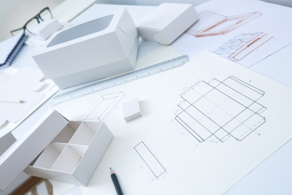 A workspace with packaging prototypes, dieline drawings, a ruler, and sketches used for designing custom packaging.